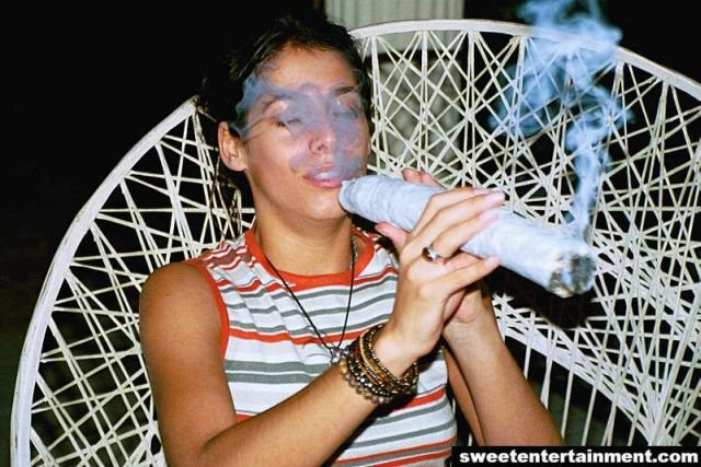 A young woman sitting in a white wicker chair. She is wearing a striped tank top and has multiple bracelets on her wrists. The woman is holding a white pipe in her right hand and is smoking it. The smoke from the pipe is coming out of her mouth. The background is dark suggesting that the photo was taken at night.