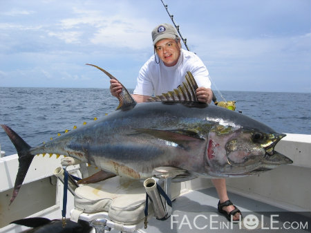 A man on a boat holding a large fish. The man is wearing a white t-shirt a cap and sunglasses and is smiling at the camera. He is holding the fish with both hands and appears to be proud of it. The fish is a yellowfin tuna which is a type of fish commonly caught in the ocean. The background shows the ocean and the sky is blue with some clouds. The boat is white and there are other fishing equipment visible in the background.