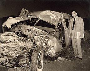 A black and white photograph of a man standing next to a car that has been severely damaged. The car appears to be a vintage model with a large front wheel and a smaller rear wheel. The hood of the car is crumpled and the windshield is shattered. The man is wearing a suit and tie and is looking at the camera with a serious expression. The background is dark suggesting that the photo was taken at night.