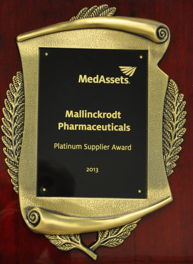 Of a gold-colored plaque with a black background. The plaque is in the shape of a shield with a curved top and a laurel wreath design on either side. The text on the plaque reads "MedAssets Mallinckrodt Pharmaceuticals Platinum Supplier Award 2013". The plaque appears to be mounted on a wooden surface.