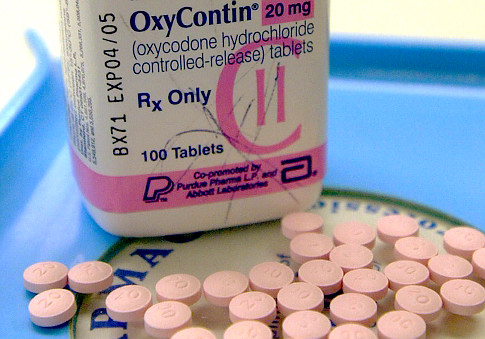 A white bottle of OxyContin 20 mg (oxycodone hydrochloride controlled-release tablets) with a pink label. The bottle is placed on a blue tray with a pile of pink pills scattered around it. The label on the bottle reads "Rx Only" and "100 Tablets". The background is blurred but it appears to be a medical setting.