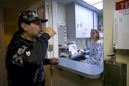 A man and a woman in a laboratory setting. The man is wearing a black baseball cap and a black jacket with a red and white logo on it. He is standing in front of a counter with a sink and a faucet. The woman is standing behind the counter and appears to be looking at the man with a concerned expression. On the right side of the image there is a sign that reads "TEST TEST" and on the left side there are several test tubes and other scientific equipment. The background shows a white wall with a window and a door.