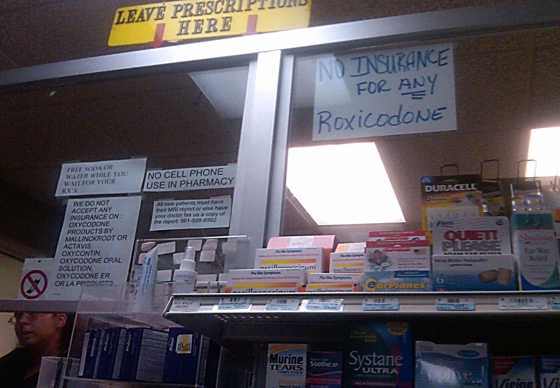 A shelf in a pharmacy with a sign above it that reads "Leave Prescriptions Here". The shelf is filled with various prescription bottles and boxes of different sizes and colors. On the left side of the shelf there is a sign that says "No insurance for any Roxicodone". On the right side there are two signs that read "No cell phone use in pharmacy" and "Duracell". The background of the image is a beige wall with a window.