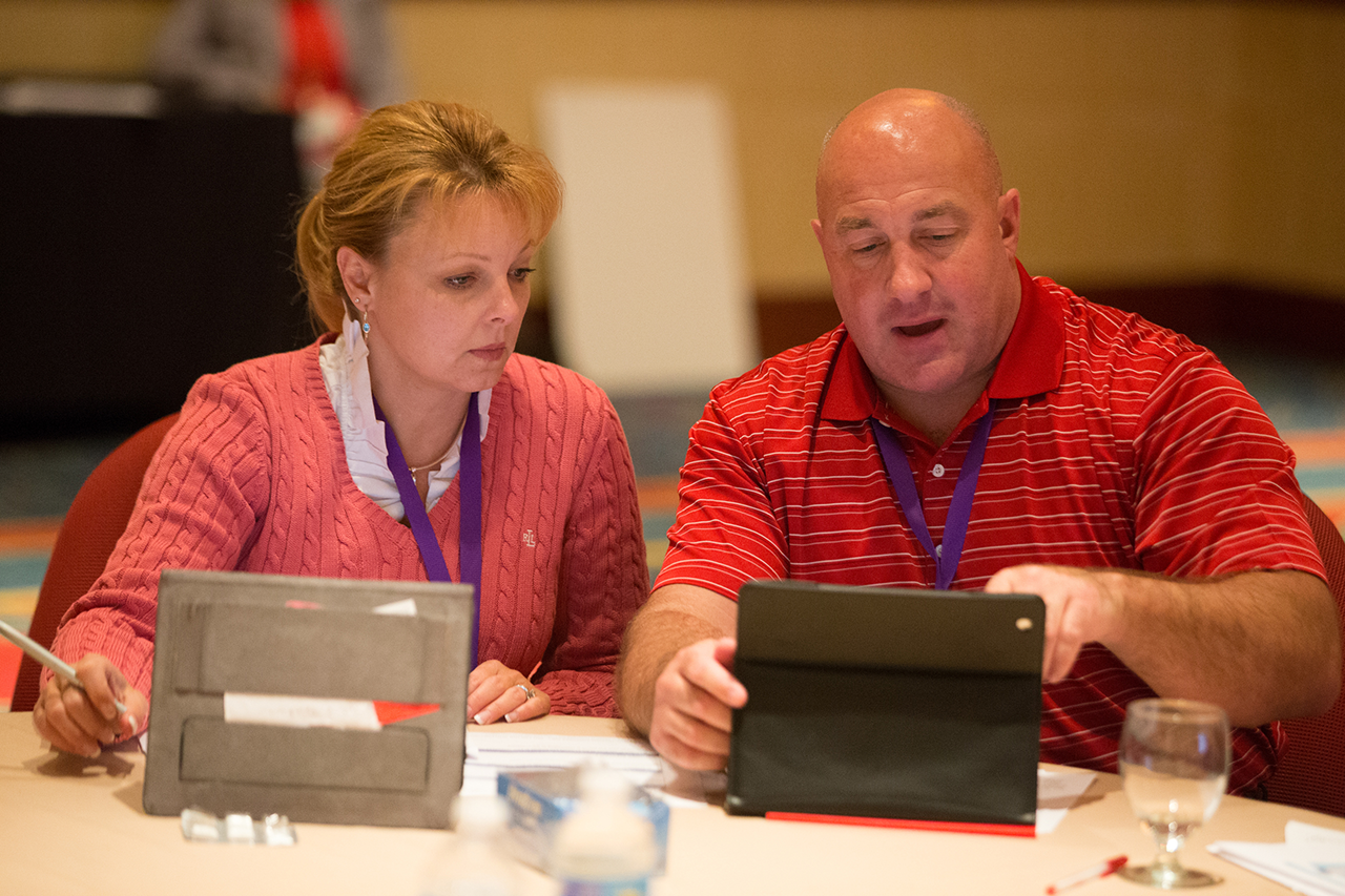 A man and a woman sitting at a table in a conference room. The man is bald and wearing a red polo shirt while the woman is wearing a pink sweater and has blonde hair. They are both looking at a tablet computer screen and appear to be engaged in a discussion. On the table there are papers a water bottle and a glass of water. The background is blurred but it appears to be a large room with a carpeted floor and a projector screen.