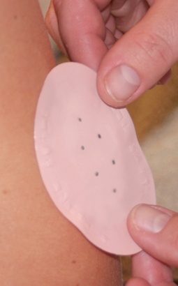 A person's hand holding a pink adhesive bandage on their arm. The bandage is oval-shaped and has small black dots on it. The person's fingers are visible on the right side of the image and they are holding the bandage with their thumb and index finger. The background is blurred but it appears to be a close-up of the person's skin.