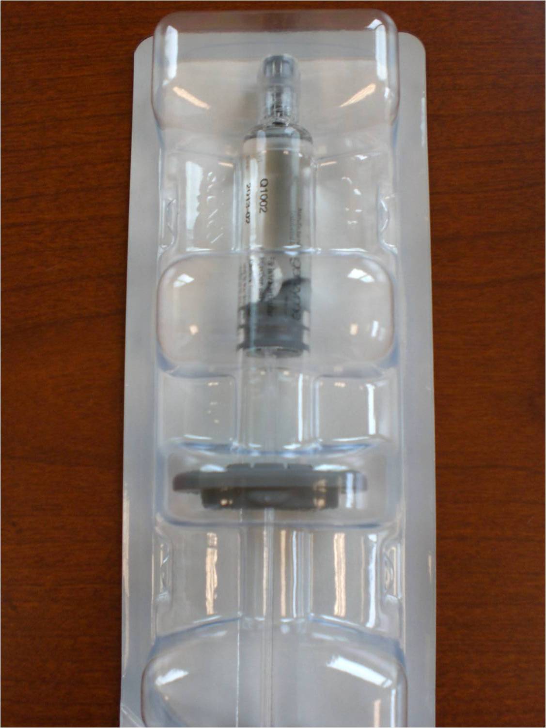 A transparent plastic packaging with a cylindrical shape. Inside the packaging there is a small metal object which appears to be a screw or a screwdriver. The screwdriver has a black handle and a silver body with a small knob on top. The body of the screwdriver is made of clear plastic and has a textured surface. The packaging is rectangular in shape and is placed on a wooden surface.