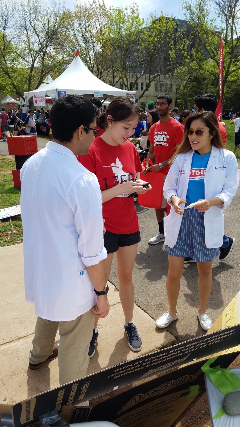 A group of people at an outdoor event. There are three people in the image two women and one man standing in front of a table with various items on it. The man is wearing a white lab coat and appears to be explaining something to the two women. The woman on the left is holding a clipboard and is looking at a piece of paper while the woman in the middle is smiling and seems to be engaged in conversation with the man on the right. In the background there are other people and tents suggesting that the event is taking place outdoors. The sky is blue and there are trees and buildings visible in the distance.