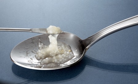 A silver spoon with a small amount of white substance likely heroin or another drug on it. A smaller metal scoop appears to be stirring or scooping the white substance from the spoon.  The spoon is resting on a blue surface.