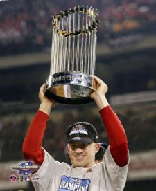 A baseball player holding up a large trophy in a stadium. The player is wearing a red and white jersey with the word "Champions" written on it and a black cap. He is smiling widely and appears to be celebrating with a big smile on his face. The trophy is silver and has multiple tiers of ribbons attached to it. The background shows a large crowd of spectators in the stands. The image appears to have been taken at night as the stadium is lit up with floodlights.