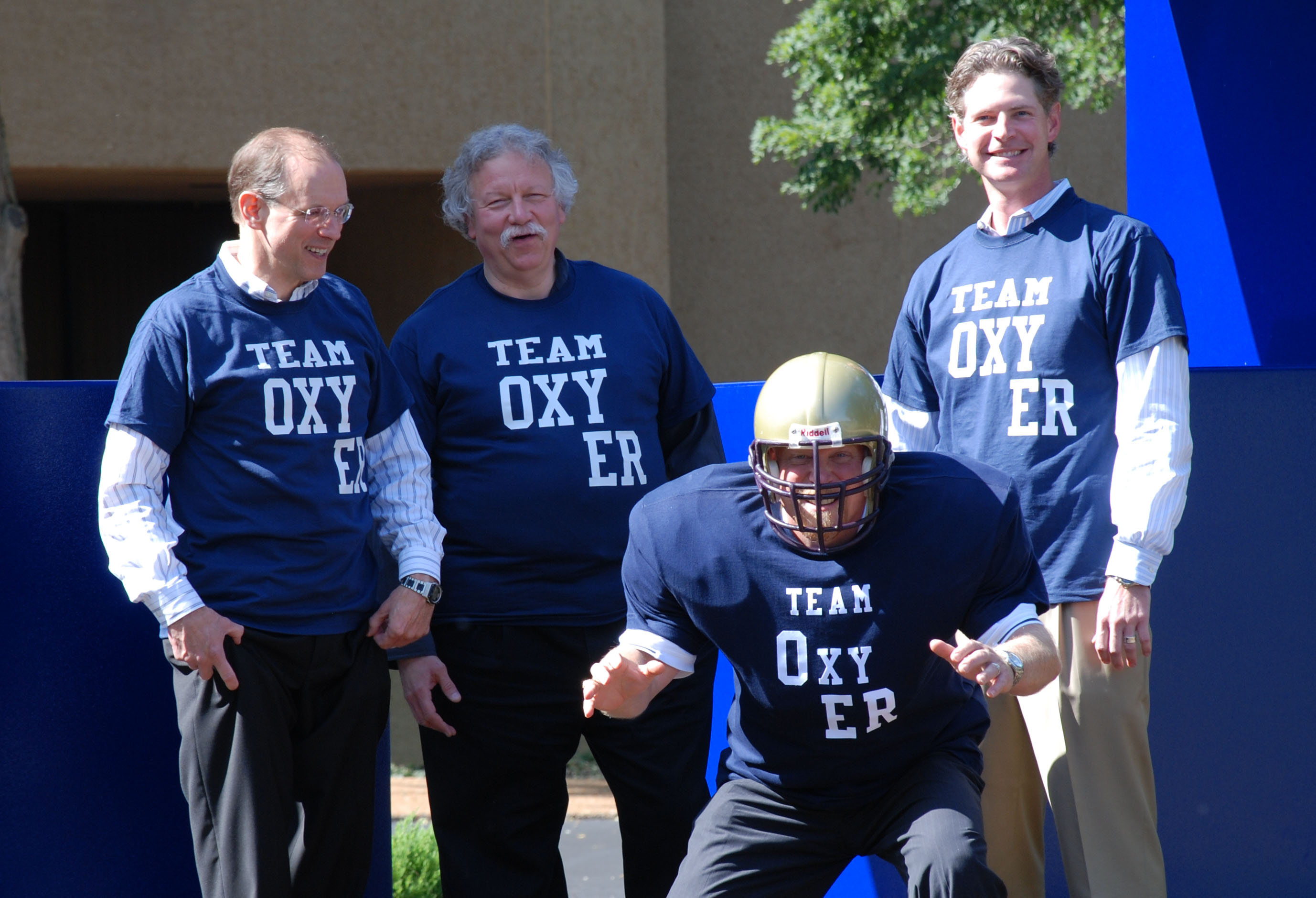 Four men standing in front of a blue wall with the words "TEAM OXY ER" written on it. They are all wearing navy blue t-shirts with the same words written in white. The man in the center is wearing a gold helmet and appears to be in the middle of throwing a football. On the left side of the image there is an older man with white hair and glasses and on the right side there are two younger men. All four men are smiling and appear to be engaged in a conversation. The background shows a building and a tree.