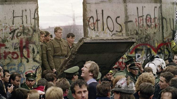 A group of people gathered around a large pile of rubble. The rubble appears to be from a collapsed building as there is debris scattered around it. The walls of the building are covered in graffiti and there are several soldiers standing on top of it looking at the rubble. Some of the soldiers are wearing helmets and uniforms while others are wearing military uniforms. In the center of the image there is a man in a suit and tie who seems to be speaking to the crowd. The sky is overcast and the overall mood of the scene is tense and chaotic.