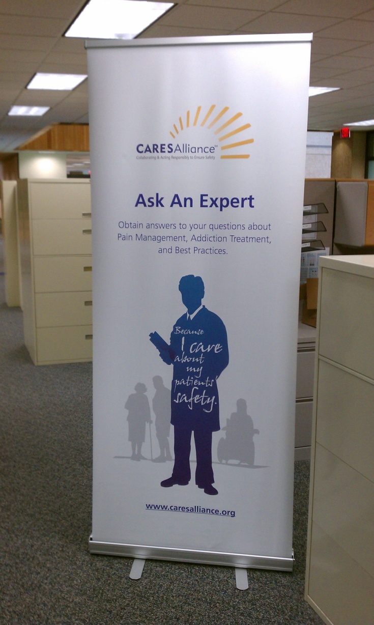 Of a roll-up banner in an office setting. The banner is white with blue text and illustrations. The text reads "Ask An Expert" and "Obtain answers to your questions about pain management addiction treatment and best practices." Below the text there is an illustration of a person holding a clipboard and standing in front of a group of people. The website "www.CARESAlliance.org" is also visible on the banner. The background of the banner is a light blue color.