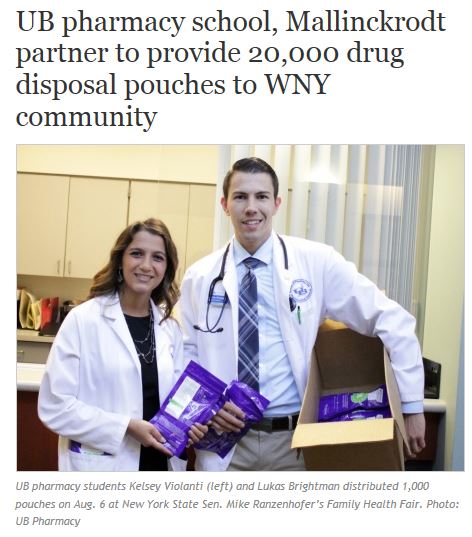 Two young people a man and a woman wearing white lab coats and stethoscopes around their necks. They are standing in a hospital room with a desk and cabinets in the background. The man is holding a cardboard box filled with purple disposable pouches which appear to be medical supplies. The woman is holding the box with both hands and is smiling at the camera. The text on the image reads "UB pharmacy students Kelsey Violanti (left) and Lukas Brightman distributed 1000 pouches on Aug. 6 at New York State Sen. Mike Ranzenhofer's Family Health Fair. Photo: UB Pharmacy."