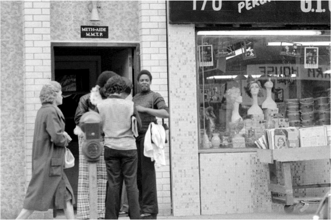 A black and white photograph of a group of people standing outside a store. The store appears to be a clothing store as there is a sign above the entrance that reads "170 Perk". The people in the image are of different ages and ethnicities and they are all looking at the store with interest.<br /><br />In the foreground there are four people two women and two men standing in front of the store. They are all wearing casual clothes and appear to be engaged in conversation. The woman on the left is wearing a long coat and carrying a bag while the man on the right is holding a jacket over his shoulder. The man in the center is wearing jeans and a t-shirt and he is looking at a display of mannequins in the window. There is also a cart with various items on it and a car parked on the street in the background.