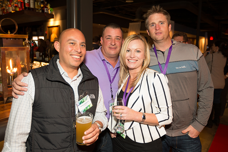 Four people standing together in a bar or restaurant. They are all smiling and holding beer glasses in their hands. The person on the left is wearing a black vest and has a name tag around his neck. Next to him there is a man wearing a purple shirt and a woman wearing a white and black striped blazer. On the right there are two men wearing striped shirts. In the background we can see other people and a bar counter with bottles of alcohol. The atmosphere appears to be relaxed and casual.