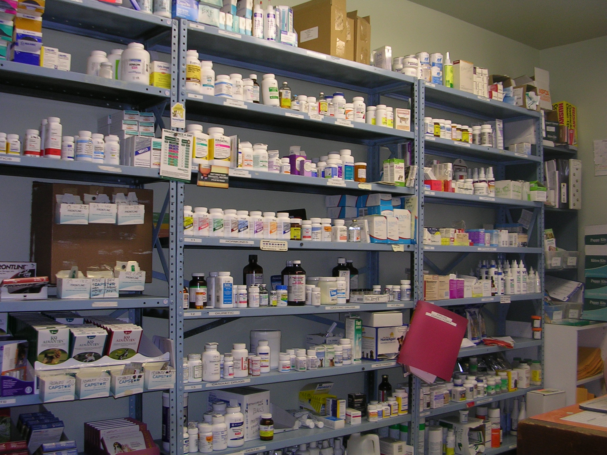 A large shelf in a pharmacy. The shelf is filled with various bottles and containers of different sizes and colors. The bottles are arranged in rows and columns with some bottles on the left side of the shelf and others on the right side. The containers are of different shapes sizes and colors and some have labels on them. There are also some boxes and other items on the shelves. The shelves appear to be well-organized and well-stocked. The background of the image is a plain white wall and there is a desk and a computer visible in the corner.