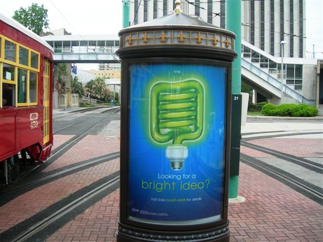 A bus stop with a large advertisement on it. The advertisement is in the shape of a lightbulb with a blue background and a green light bulb in the center. The light bulb is surrounded by a yellow border and has the words "Looking for a bright idea?" written in white text. The bus stop is located on a brick pavement with a red trolley car on the left side and a tall building in the background. The sky is overcast and the overall mood of the image is bright and cheerful.