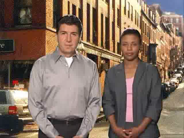 A man and a woman standing on a street in a city. The man is on the left side of the image wearing a grey collared shirt and black pants. He has short dark hair and is looking directly at the camera with a serious expression. The woman is standing next to him also wearing a gray blazer and a pink top. She has short black hair. The street is lined with brick buildings on both sides and there are cars parked on the sidewalk. The sky is blue and the weather appears to be sunny.