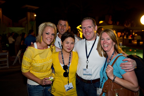 A group of five people posing for a photo at an outdoor event. There are six people in the photo three men and three women all of whom are smiling and looking at the camera. The woman on the left is wearing a yellow top and holding a drink in her hand. The man in the middle is wearing an ID card and is standing next to the woman in the yellow top. The other three people are standing behind him also smiling. In the background there are other people and tents suggesting that the event is taking place outdoors at night.