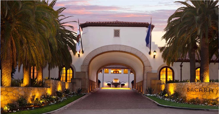 The entrance of a luxurious hotel named Bacara. The entrance is a large archway with a tiled roof and arched windows. The archway is surrounded by palm trees and other tropical plants. The hotel is lit up with warm yellow lights creating a warm and inviting atmosphere. The sky is a beautiful orange and pink color indicating that it is either sunrise or sunset. The word "Bacara" is written on the wall on the right side of the archway. The overall mood of the image is peaceful and inviting.