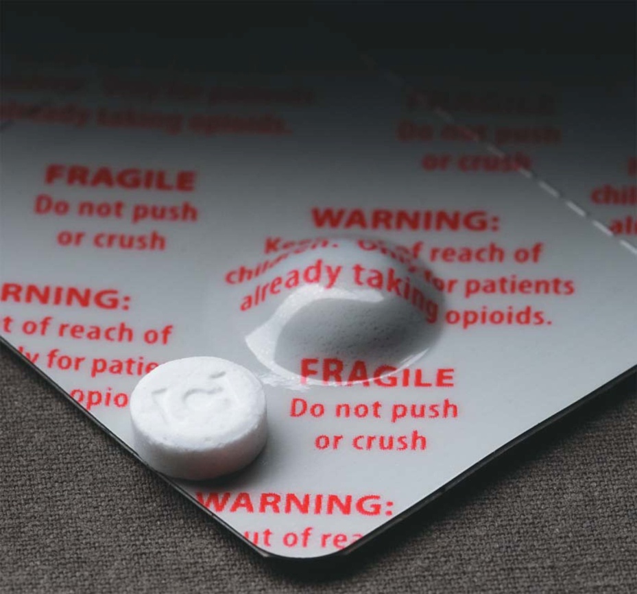 A close-up of a white pill with a red label on it. The label has the words "WARNING: Do not push or crush" written in bold black letters. Below the text there is a smaller text that reads "FRAGILE: Keep out of reach of patients already taking opioids." The label also has a small illustration of a pill on the bottom right corner. The background is a plain white surface.