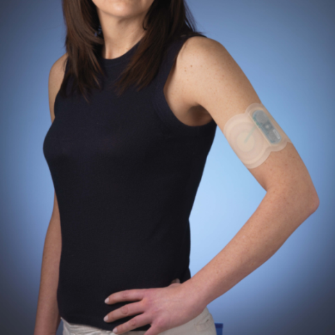 A young woman with shoulder-length brown hair wearing a black tank top and beige pants. She is standing with her left hand on her hip and her right hand resting on her thigh. On her right arm there is a white bandage with a blue label on it. The bandage appears to be a medical device possibly a bandage or a gauze. The background is a solid blue color. The woman is looking directly at the camera with a slight smile on her face.