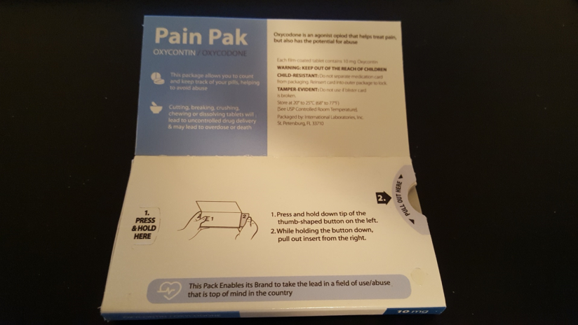 A photograph of the back of a box of Pain Pak. The box is white with blue text and illustrations on it. The top of the box has the brand name "Pain Pak" written in bold letters at the top followed by the product name "Oxycodone" in smaller letters. Below that there is an illustration of a hand holding a pain pack. <br /><br />On the right side of the image there are instructions on how to use the pain pack including pressing the button on the left side pressing the right button and pressing the bottom button. The instructions also mention that the pack enables the brand to take the lead in a field of use.<br /><br />The box is placed on a black surface and the background is blurred.