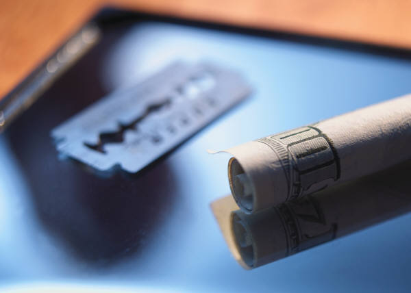 A close-up of a razor blade and a roll of toilet paper on a blue surface. The razor blade is on the left side of the image with the blade facing towards the right side. The roll of paper is rolled up and appears to be empty. The background is blurred but it seems to be a wooden table or countertop. The image is taken from a slightly elevated angle looking down on the razor blade.