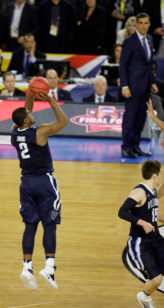 A basketball game in progress. The player in the foreground is wearing a navy blue jersey with the number 2 on it and white shorts. He is in mid-air with his arms stretched upwards and a red basketball in his hands. He appears to be in the process of taking a shot. In the background there are spectators in the stands and a referee on the sidelines. On the right side of the image there is another player from the opposing team wearing a white jersey with blue stripes. The image appears to have been taken from the sidelines of the court.