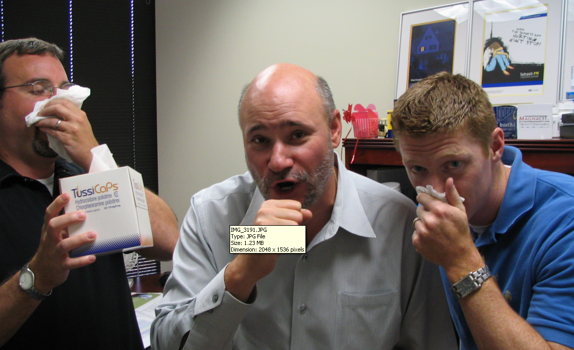 Three men in an office setting. The man in the middle is bald and has a surprised expression on his face. He is holding a box of TussiCaps which is white and blue in color. The other two men are also holding tissues and appear to be blowing their noses. One of the men on the left is wearing a black shirt and glasses while the other two are wearing blue shirts. In the background there is a desk with a computer and other office supplies.