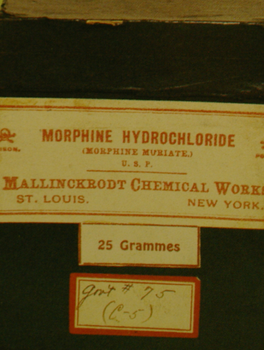 A close-up of a label on a black box. The label is rectangular in shape and has a red border. The text on the label reads "Morphine Hydrochloride" in bold white letters. Below the text there is a smaller text that reads "U.S.P. MALLINKRODT CHEMICAL WORKS ST. LOUIS NEW YORK 25 Grammes". The label also has a small red square with the number "25 Grammes" written in white letters at the bottom. The background of the label is black.