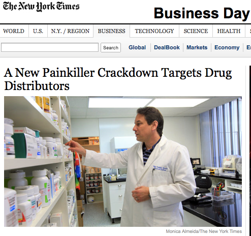 A man in a white lab coat standing in a pharmacy. He is pointing to a shelf with various bottles and containers on it. The man appears to be a pharmacist or a drug distributor as he is wearing a lab coat and has a serious expression on his face. The background shows a countertop with a computer and other medical equipment. The image is accompanied by text that reads "A New Painkiller Crackdown Targets Drug Distributors" and "The New York Times Business Day".