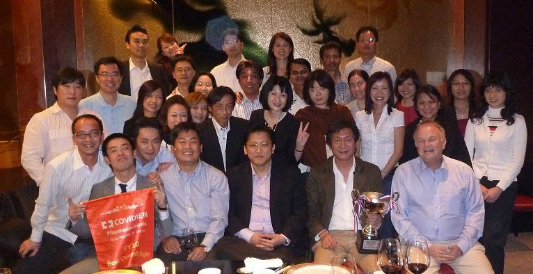 A group of people posing for a group photo in a restaurant. There are around 20 people in the photo all of them are smiling and looking at the camera. In the center of the group there is a man holding a red banner that reads "Covidien" and a trophy. The group is sitting at a table with plates of food and glasses of wine. The background appears to be a large room with a chandelier hanging from the ceiling. The overall mood of the photo is celebratory and happy.