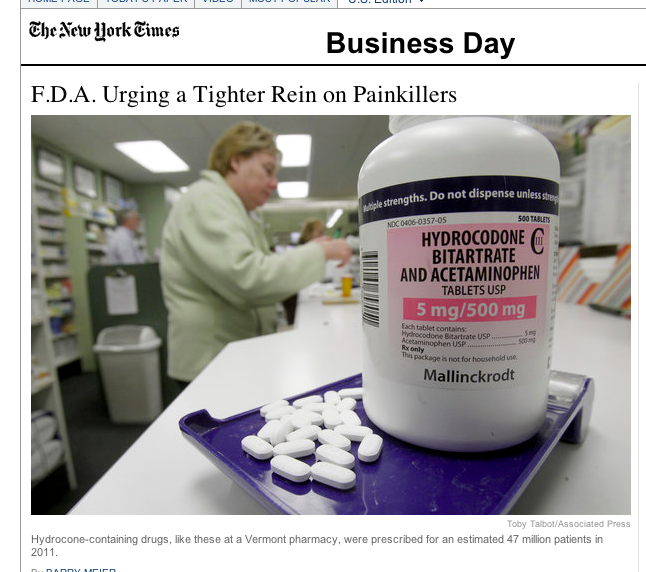 A white bottle of Hydrocodone Bitrate and Acetaminophen tablets on a purple tray. The bottle has a label that reads "5 mg/500 mg" and "Mallinckrodt". In the background there is a woman in a green lab coat working in a pharmacy. The image appears to be a screenshot from a news article about the company's business day.