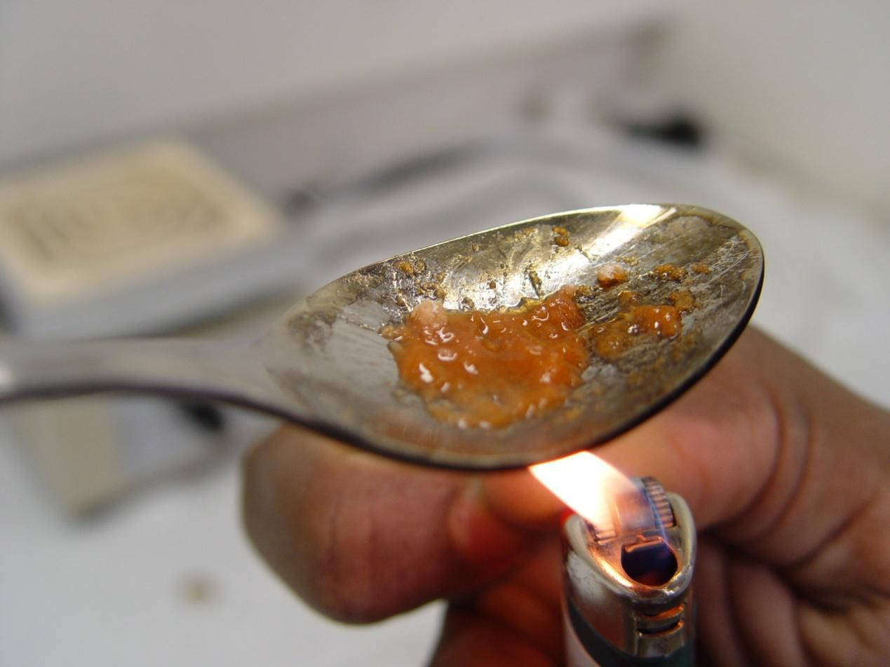 A silver spoon with a small amount of orange-colored substance on it likely heroin or another drug. A person's hand is holding a lit lighter under the spoon. The background is blurred but it seems to be a kitchen countertop.