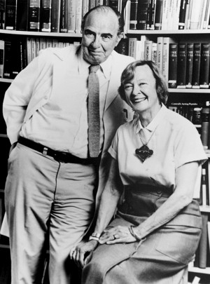 A black and white photograph of an elderly couple posing for a photo in front of a bookshelf. The man is on the left side of the image wearing a suit and tie and the woman is sitting on the right side. They are both smiling and looking at the camera. The woman is wearing a short-sleeved blouse and a necklace with a pendant. The bookshelves behind them are filled with books of various sizes and colors. The image appears to be from the early 20th century.