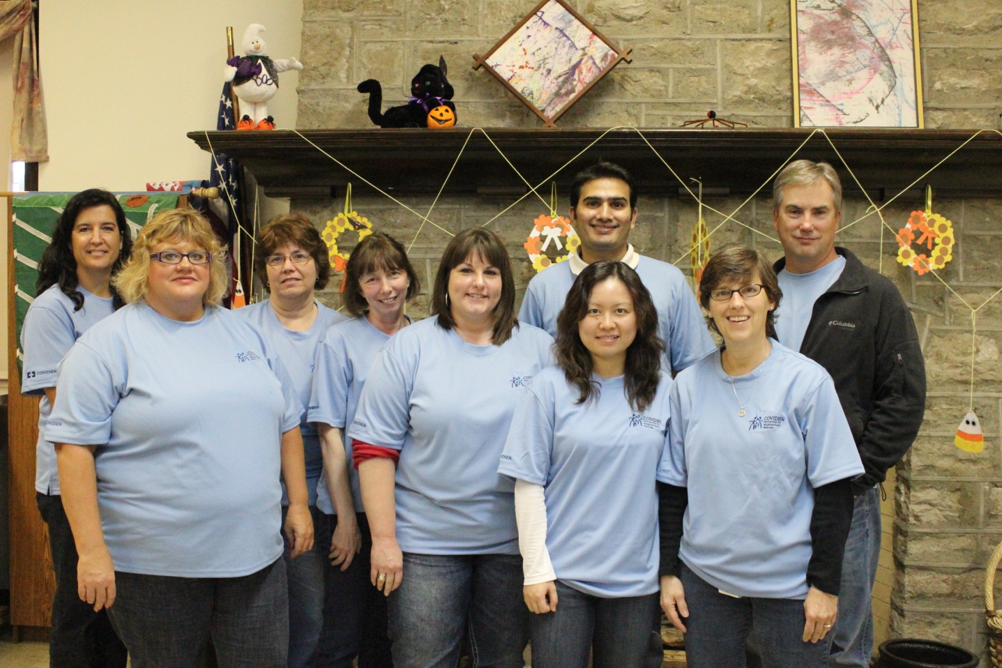 A group of nine people standing in front of a fireplace. They are all wearing light blue t-shirts and are smiling at the camera. The fireplace is decorated with various items such as a stuffed animal a painting and a picture frame. There are also some decorative items hanging on the wall behind the fireplace. The people in the group appear to be of different ages and ethnicities.