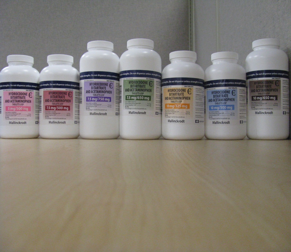 A row of nine white plastic bottles arranged in a line on a wooden surface. The bottles are of different sizes and shapes with the largest bottle at the bottom and the smallest at the top. Each bottle has a label on it with a different color and text. The labels appear to be for different types of supplements or supplements. The background is a plain white wall.