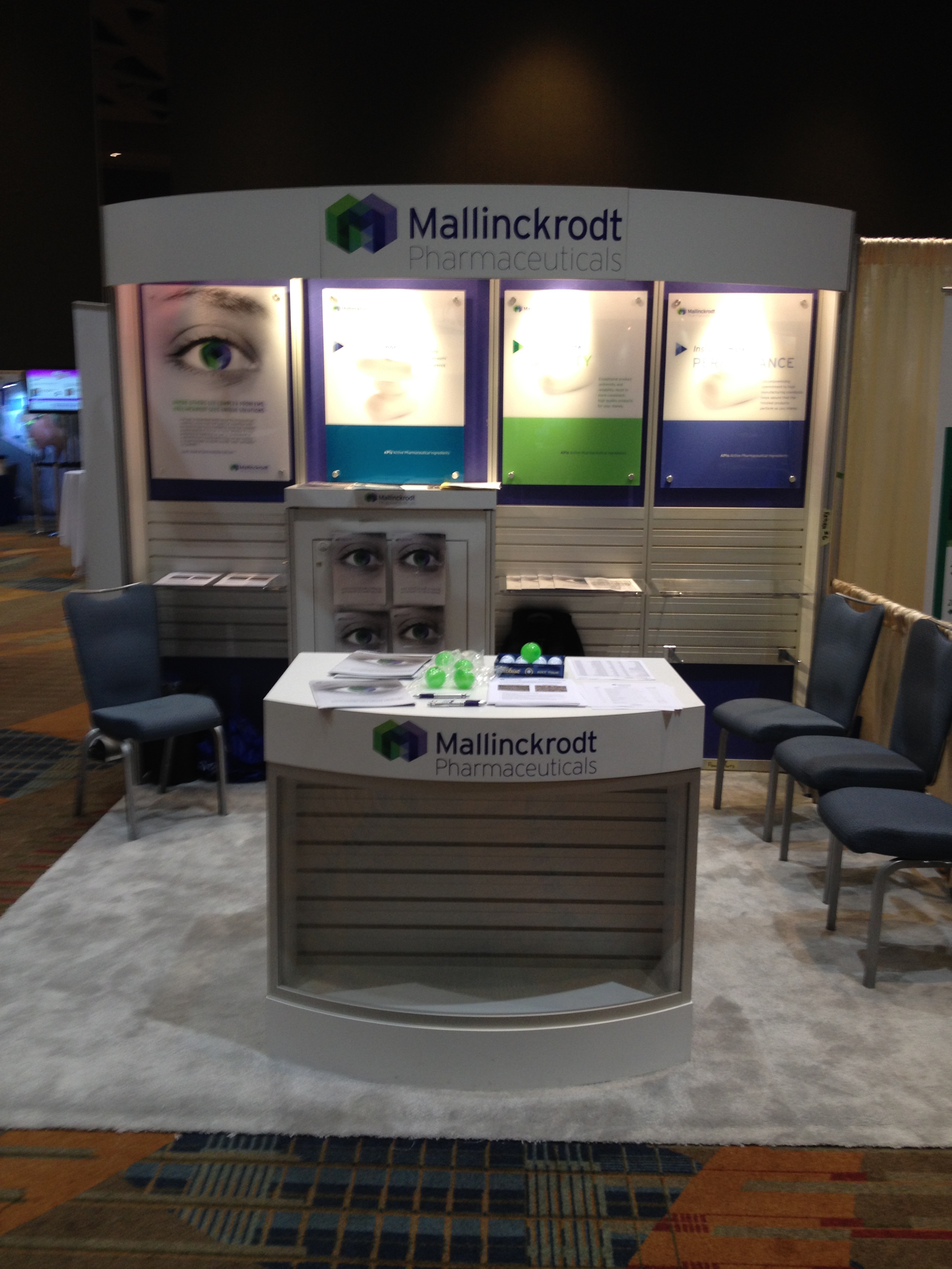 A booth at a trade show or exhibition. The booth is white with a curved design and has a sign that reads "Mallinckrodt Pharmaceuticals". There are three large screens on the wall behind the booth each displaying an image of a person's face. On the right side of the booth there are four blue chairs arranged in a semi-circle and on the left side there is a desk with a computer monitor and other items on it. The floor is covered with a colorful patterned rug. The background is dark and there are other booths visible in the background.