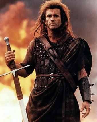 A still from a movie or TV show. It shows a man with long curly hair and a serious expression on his face. He is holding a sword in his right hand and appears to be ready for battle. The man is wearing a dark-colored tunic with a high collar and a belt with spikes on it. The tunic is draped over his shoulders and he is also holding a shield in his left hand. The background is a fiery orange and yellow color suggesting that the scene is taking place at night. The overall mood of the image is intense and dramatic.