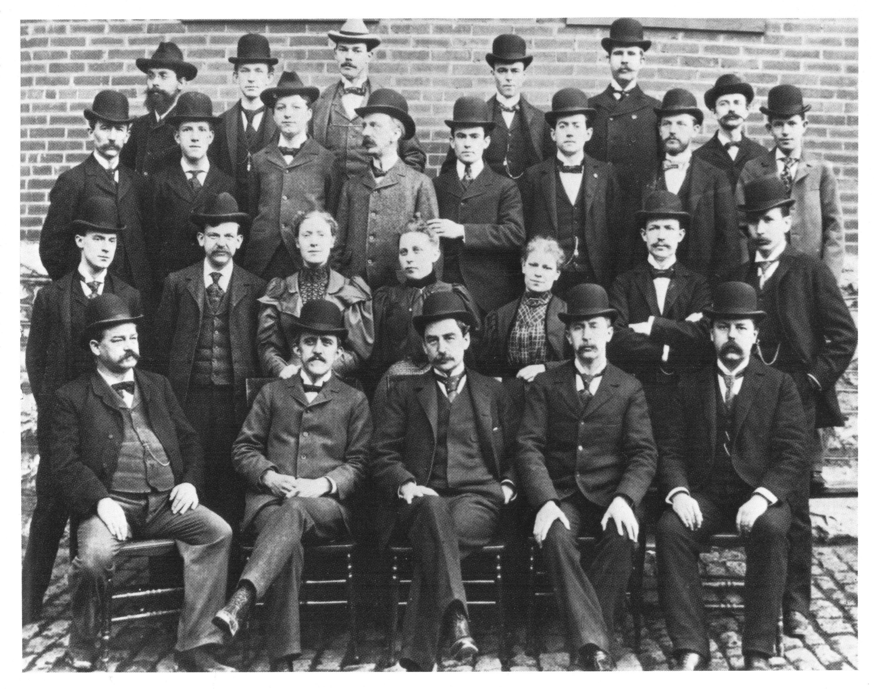 A black and white photograph of a large group of men and women. They are all dressed in formal attire with some wearing hats and others wearing suits. The men are arranged in a semi-circle facing the camera and they are all facing the same direction. The background appears to be a brick wall and the photograph is taken from a low angle looking up at the group. The overall mood of the image is serious and professional.