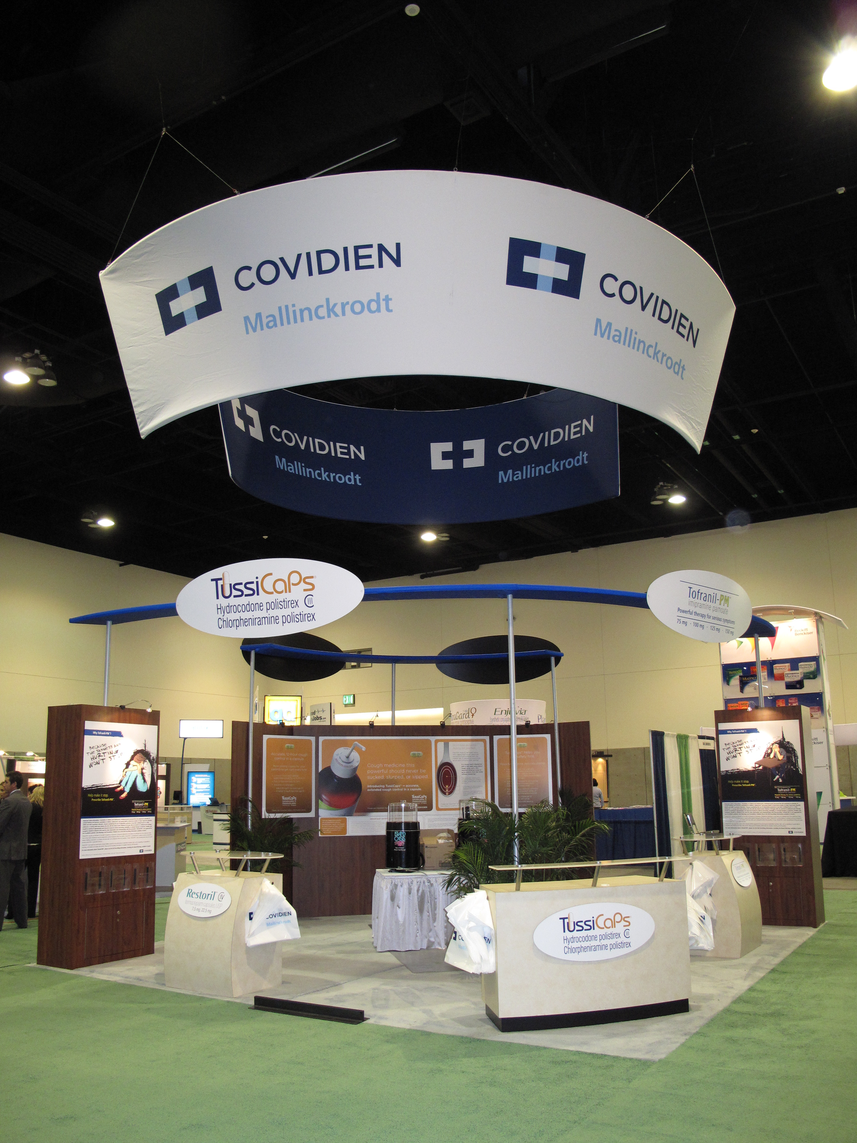 A trade show booth with a large white banner hanging from the ceiling. The banner has the logo of Covidien Mallinckrodt and the words "Covidien" written in blue. Below the banner there is a sign that reads "TussiCaps" in white letters. The booth is set up on a green carpeted floor and there are several other booths and displays in the background. There are also a few people walking around the booth. The overall atmosphere of the booth is modern and professional.