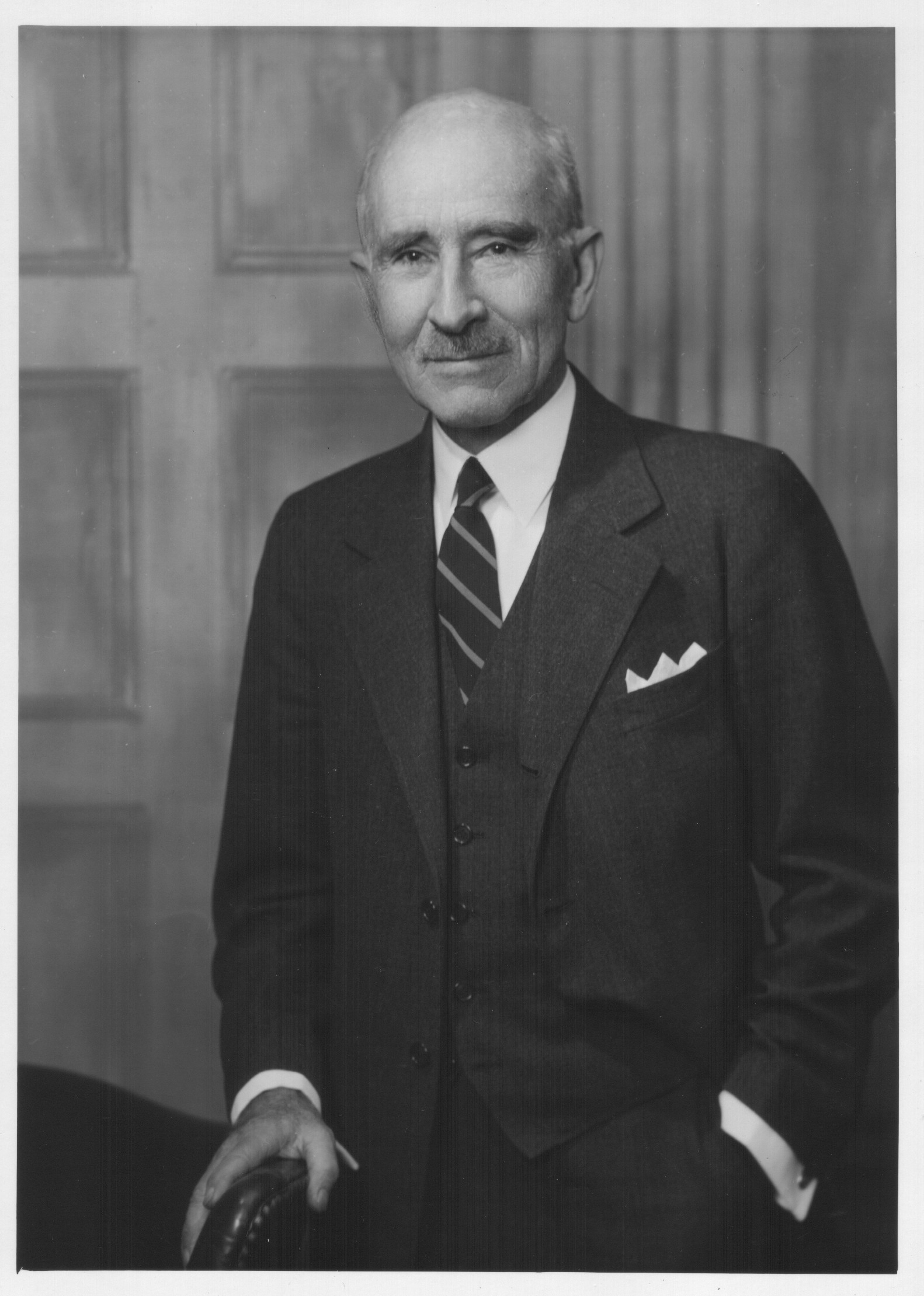 A black and white portrait of a middle-aged man. He is wearing a formal suit with a striped tie and a white pocket square. He has a serious expression on his face and is looking directly at the camera. The background appears to be a room with a wooden paneled wall and a chair. The man is standing with his left hand resting on the armrest of the chair.