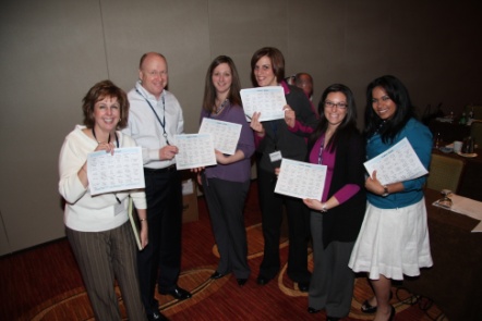 A group of six people standing in a room with a carpeted floor and a wall in the background. They are all holding up certificates in their hands and are smiling at the camera. The people appear to be of different ages and ethnicities and they are dressed in formal attire. The certificates they are holding are white with black text and have a list of names and dates printed on them. There are also a few other people in the room but they are not visible in the image.