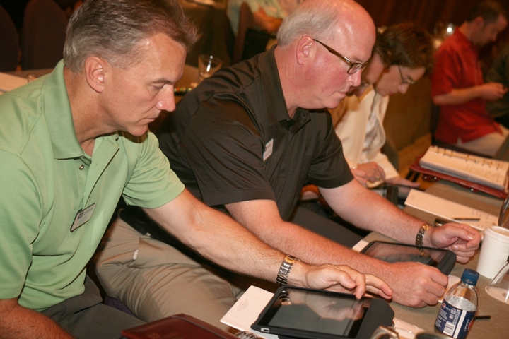 Two older men sitting at a table and using a tablet computer. They are both wearing green polo shirts and appear to be focused on the screen of the tablet. The man on the left is holding the tablet in his hands and appears to be using it while the other man is looking at it intently. There are other people sitting at the table in the background some of whom are also using their phones. The table is covered with a white tablecloth and there are various items on it including a water bottle a coffee cup and some papers. It appears that they are at a conference or event.