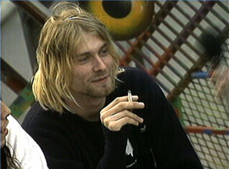 A young man with long blonde hair and a beard sitting in a room with a colorful background. He is holding a cigarette in his right hand and appears to be deep in thought. He has a serious expression on his face and is looking off to the side. The man is wearing a black sweatshirt and there is another person visible on the left side of the image.