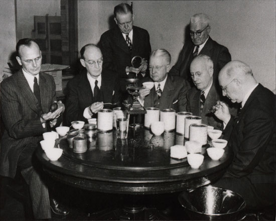 A black and white photograph of a group of six men sitting around a round table. They are all wearing suits and appear to be engaged in a conversation. The table is covered with a tablecloth and there are several cups and saucers on it. The men are of different ages and ethnicities and they are all looking at the cups with serious expressions on their faces. One of the men is holding a cup of tea or coffee while the others are looking down at the table. In the background there is a man standing and holding a glass of water. The photograph appears to be from the early 20th century.