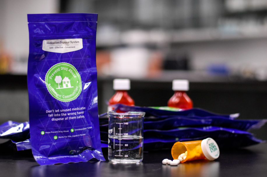 A purple packet with a green label on it. The label has a picture of a house and the text "Don't let unused medications fall into the wrong hand. Dispose of them safely." Next to the packet there is a small glass beaker filled with water and a small orange pill bottle with a white cap. The background is blurred but it appears to be a laboratory.