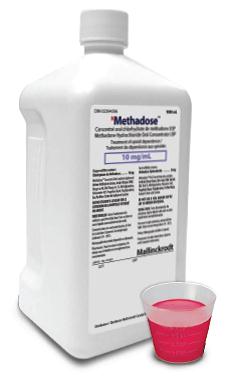 A white plastic jug with a white cap and a label on it. The label has the brand name "Methadose" written in bold black letters at the top followed by the product name "10 mg/ml" in smaller black letters. Below the label there is a list of ingredients and their corresponding nutritional information. Next to the jug there are two small plastic cups one filled with a pink liquid and the other with a red liquid. The background is plain white.