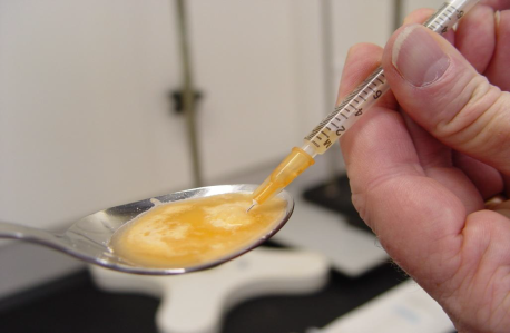 A hand holding a syringe with a needle inserted into a spoon. The syringe is filled with a yellow liquid which appears to be a mixture of orange-colored liquid and the spoon is resting on a white countertop. The background is blurred but it seems like the person is in a kitchen or a laboratory setting.